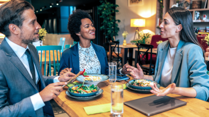 Tre personer i formella kläder sitter vid ett bord på en restaurang och pratar medan de äter. Det finns olika maträtter och glas med vatten.