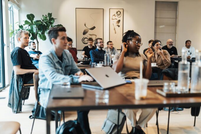 Personer sitter i ett ljust rum med laptops och antecknar, lyssnar uppmärksamt. Gröna växter och abstrakt konst på väggarna.